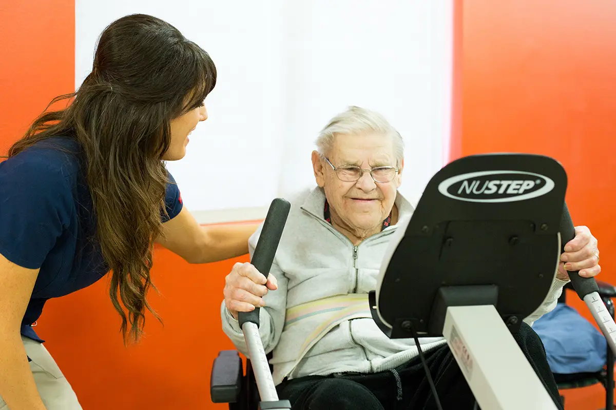Elderly woman using cardiac rehabilitation exercise equipment with staff guidance