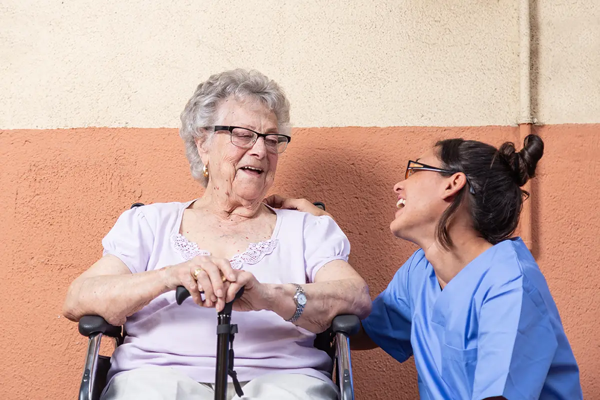 Elderly woman with glasses smiling while receiving attentive care from a staff member
