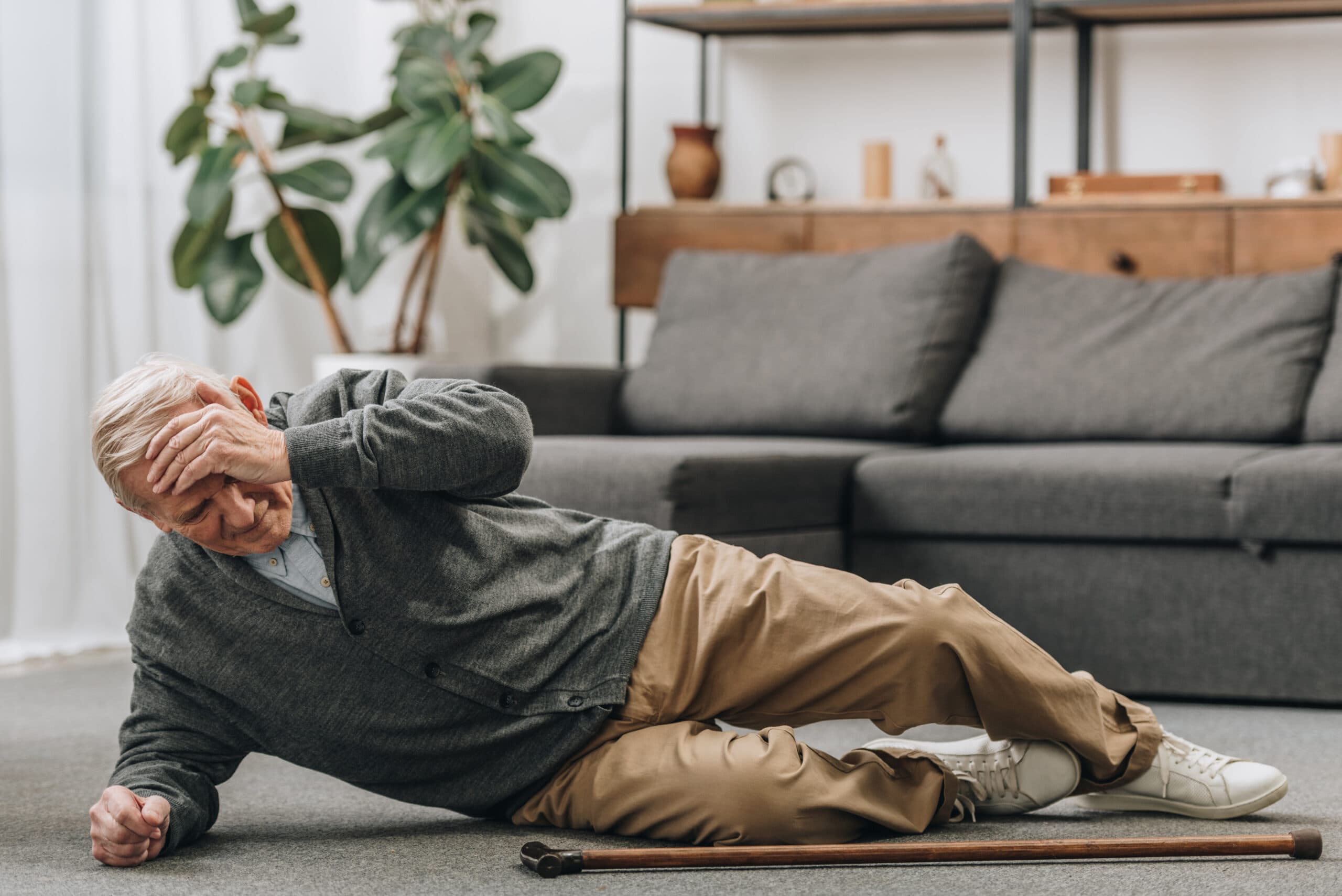 A senior man lying on the floor with his cane next to him holding his head in discomfort