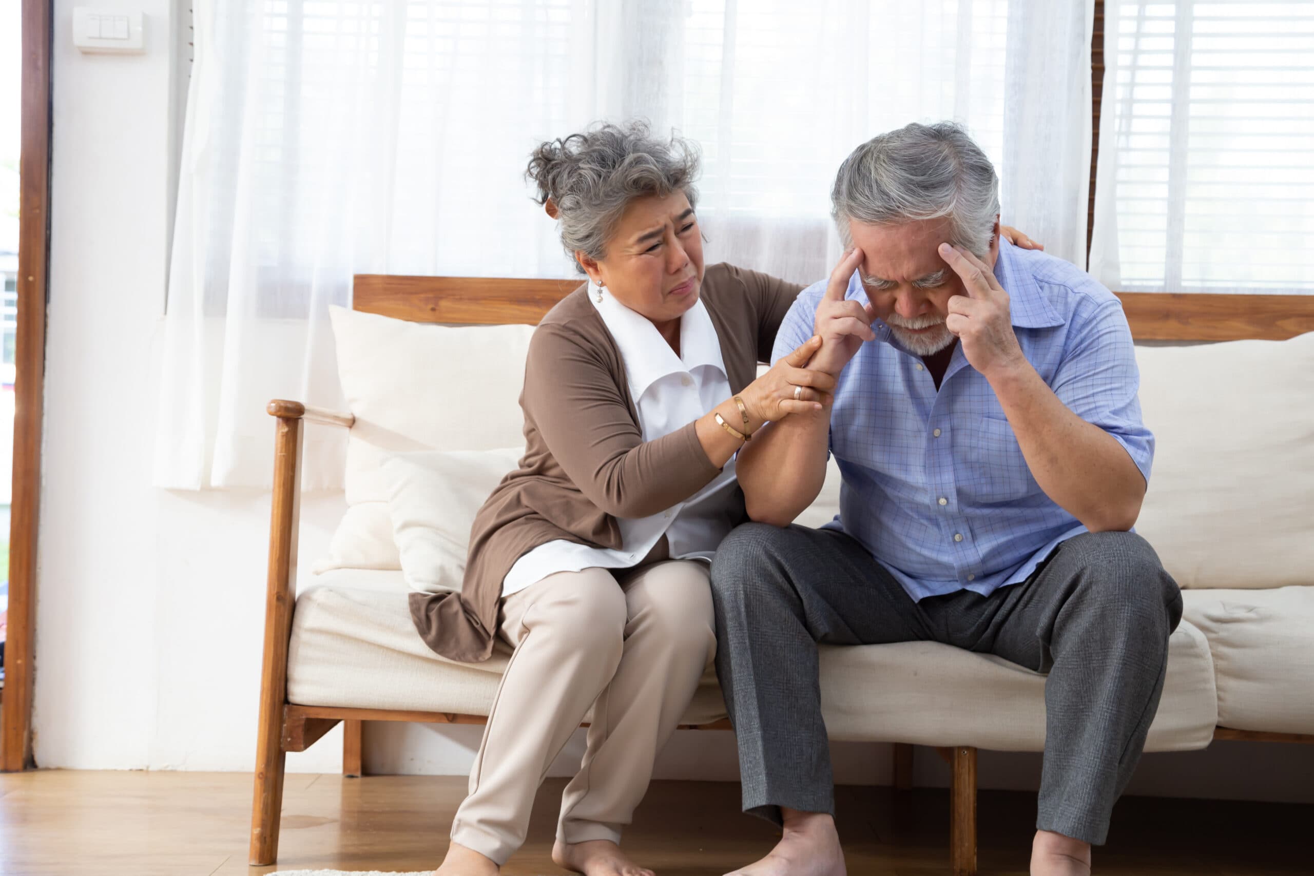 Older man sitting on a couch holding his head while a woman comforts him