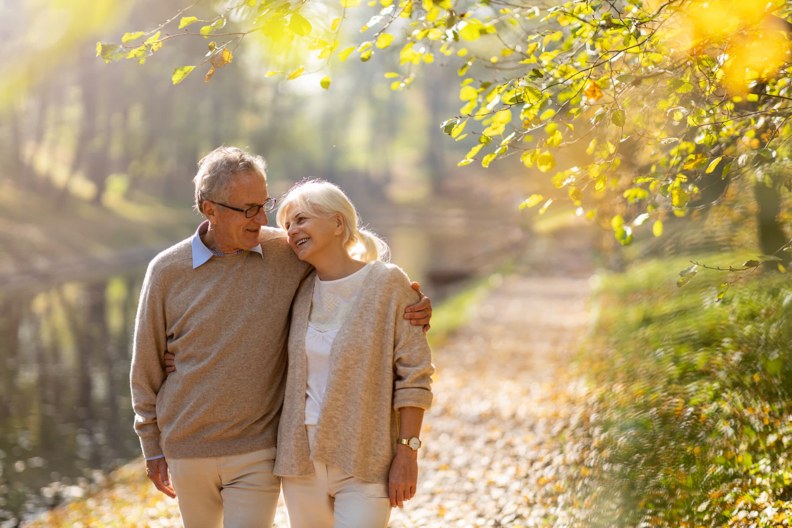 An image of a senior man with his arm around a senior woman walking down a sunny, fall path